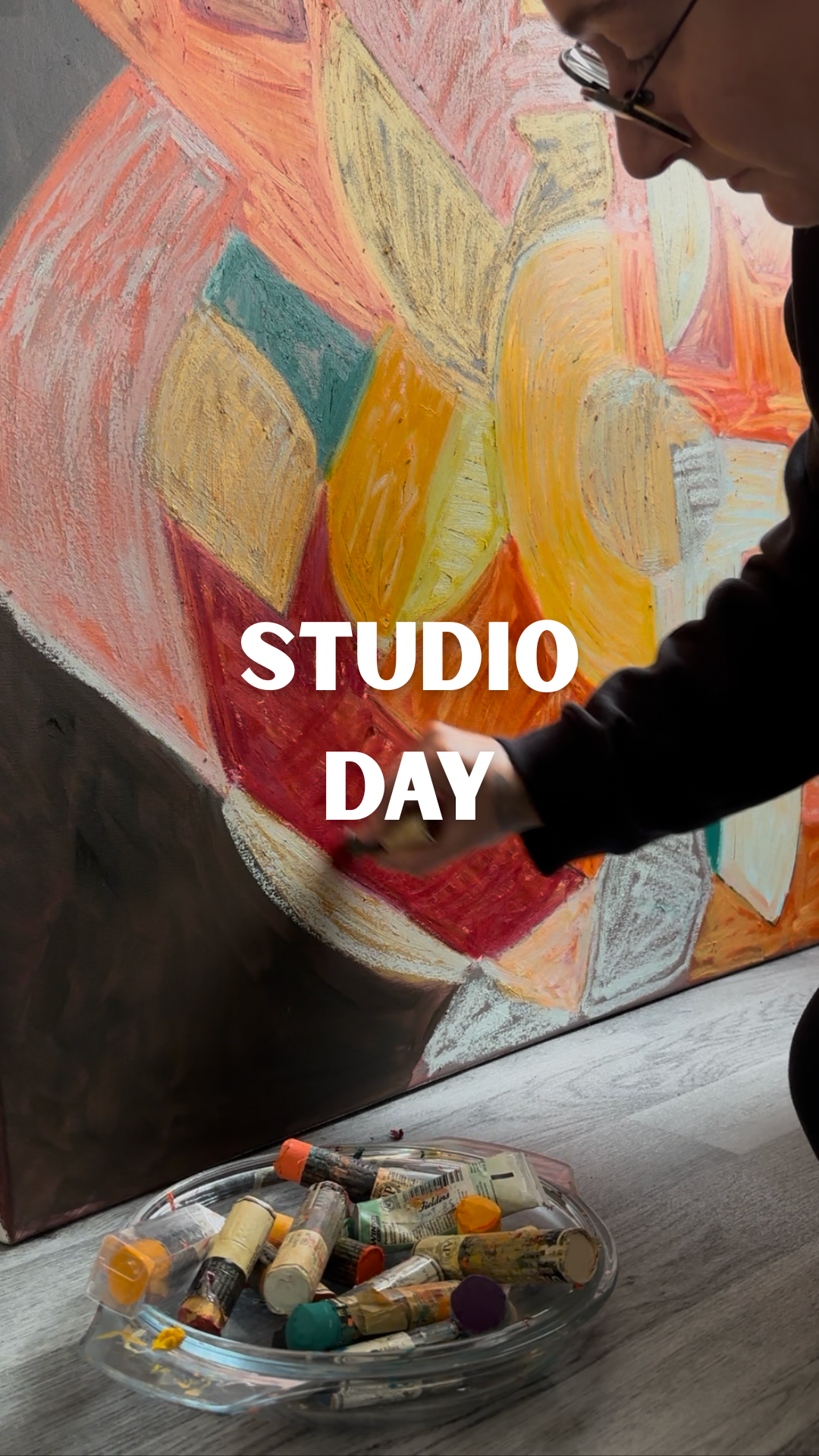 Anastasija Sarajeva working with an oil stick on a large textured abstract painting in coral, yellow, cream and teal, with art materials below. Text reads: “Studio day.”