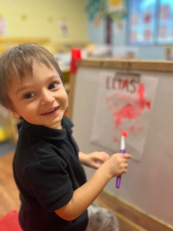A young boy smiling and holding a marker in front of a whiteboard with red writing and drawings in a classroom.