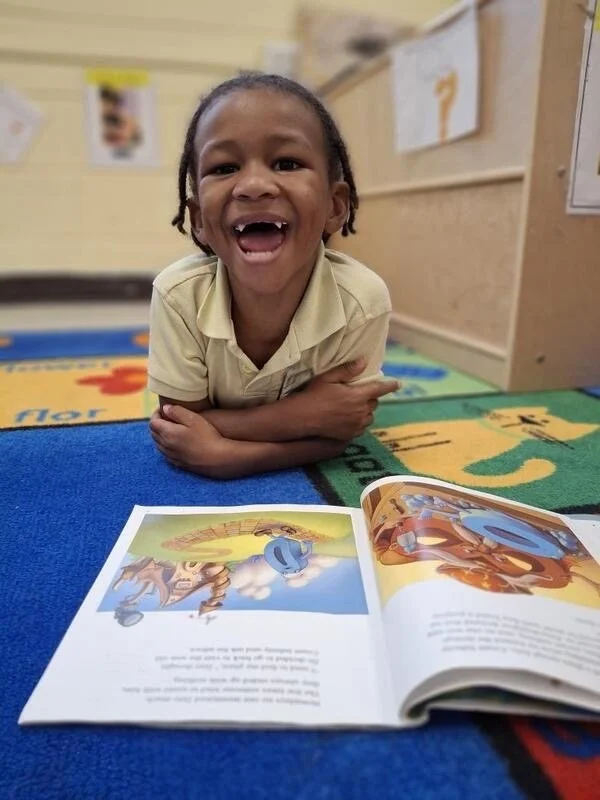 A young child lying on the floor, smiling broadly, with an open children's book in front of them on a colorful carpet at a preschool or classroom setting.