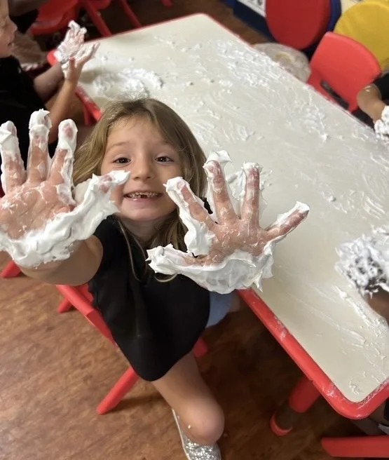 A young girl with a big smile showing her hands covered in white foam, sitting at a table in a classroom or play area.