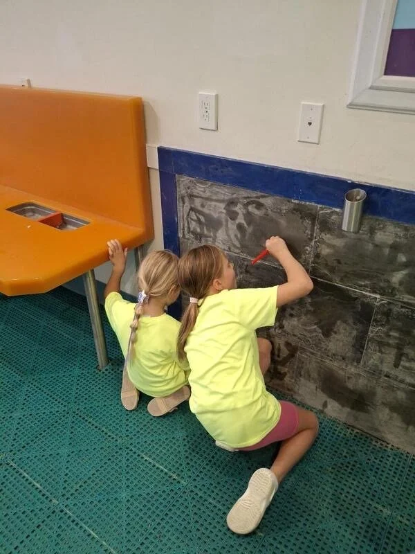 Two girls in yellow shirts drawing on a wall with a red marker in a hospital or clinic room with a blue-tiled lower wall and orange furniture.
