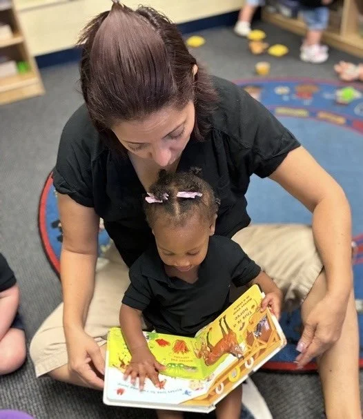 An adult woman reading a colorful children's book to a young girl sitting on her lap in a library or classroom setting.