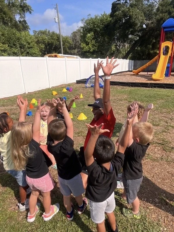 Group of children celebrating outdoors with their hands raised in front of a playground slide and scattered cones.