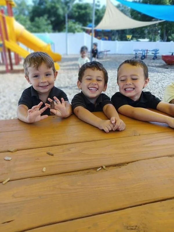 Three young boys sitting at a wooden picnic table outdoors, smiling and enjoying themselves. In the background, there is a playground slide, colorful shade sails, and other children playing.