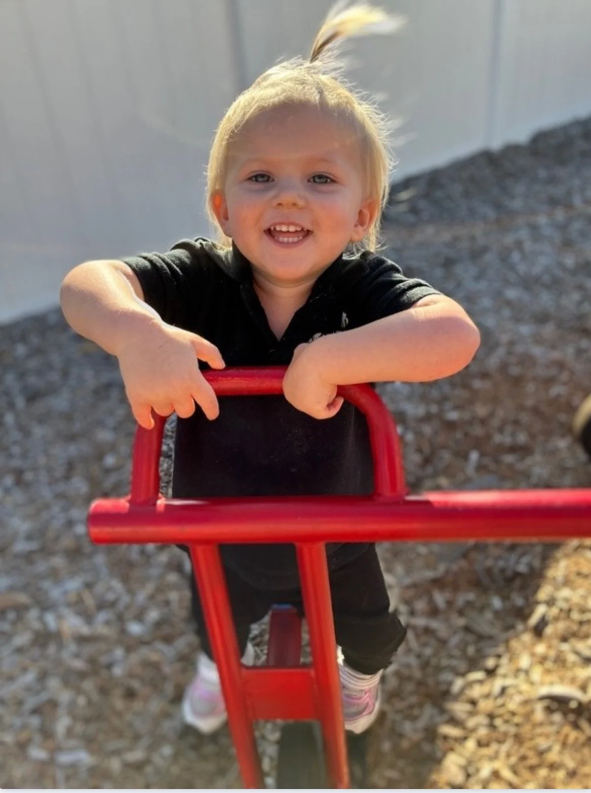 A young girl with blonde hair smiling, holding onto a red skateboard, standing on a gravel surface outdoors.