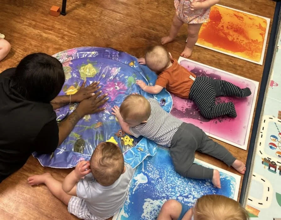 Children and an adult engaging in a sensory play activity with colorful textured mats and a splash mat on a wooden floor.