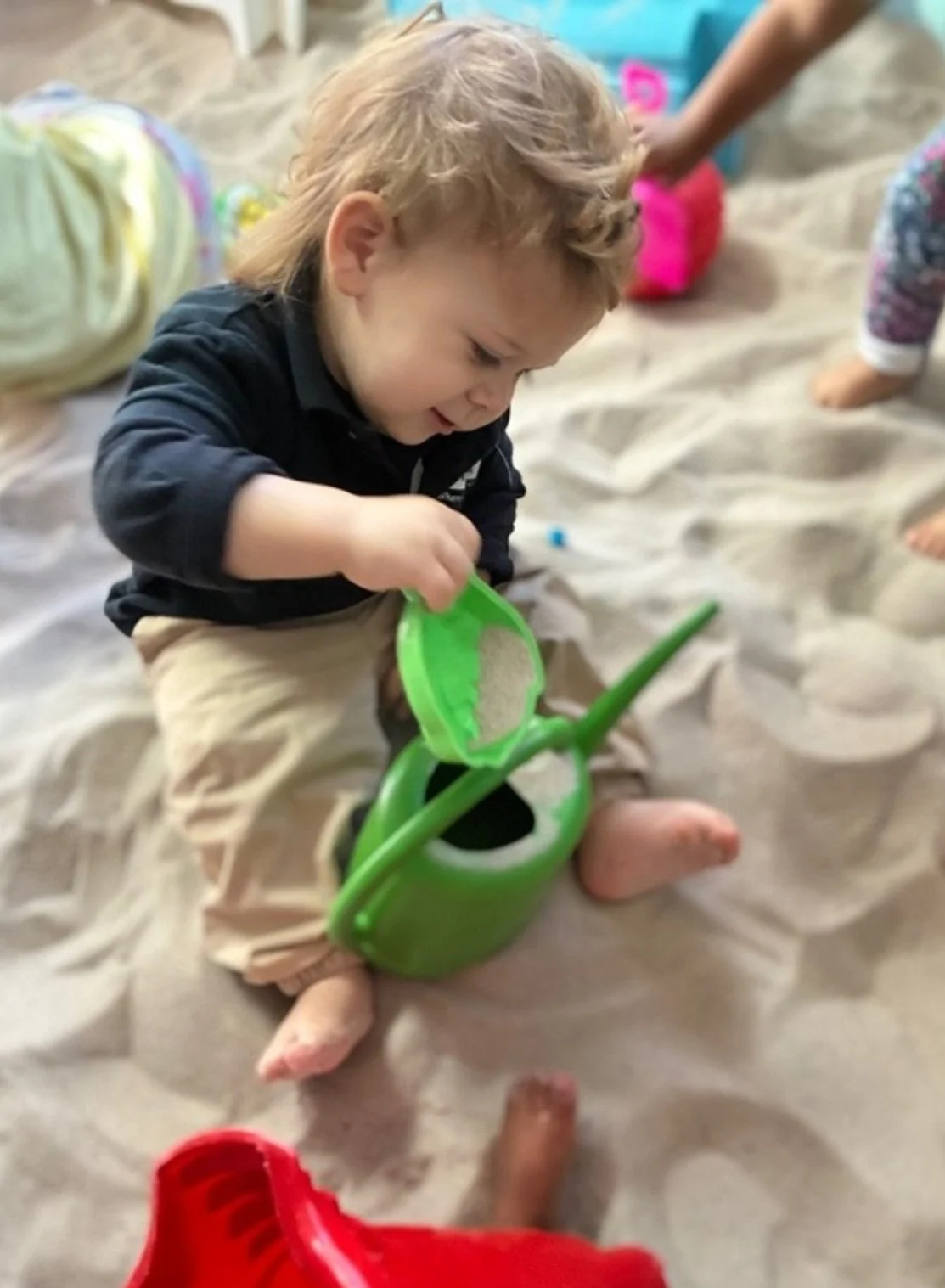 Young child sitting on sandy floor, playing with a green watering can and a green sand shovel, surrounded by other children and beach toys.
