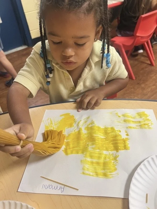 A young girl with braided hair and blue beads is painting yellow streaks on a white sheet of paper with a paintbrush. She appears focused on her art project in a classroom setting.