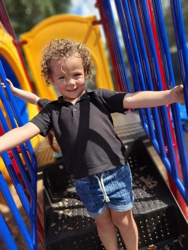 A young child with curly hair smiling and playing at a colorful playground.