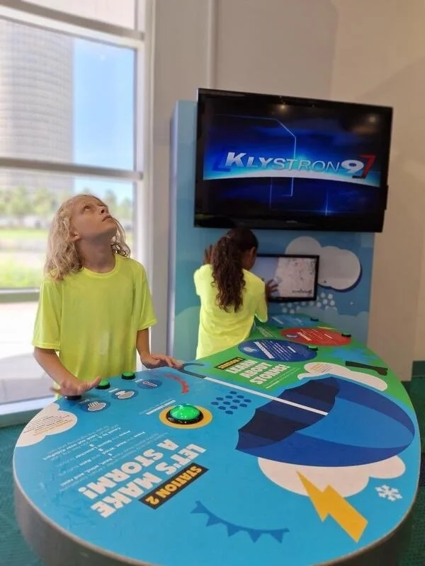 Two children in neon yellow shirts playing an interactive display at a science museum. One with curly blonde hair looks up, while the other with long brown hair interacts with a touchscreen. The display features science-themed graphics and a large screen displaying 'KY STRONG 9'.
