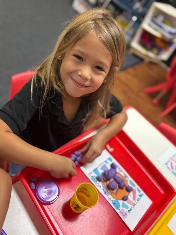 A young girl with long blonde hair smiles at the camera while sitting at a red table with a coloring page and glue stick, in a classroom setting.