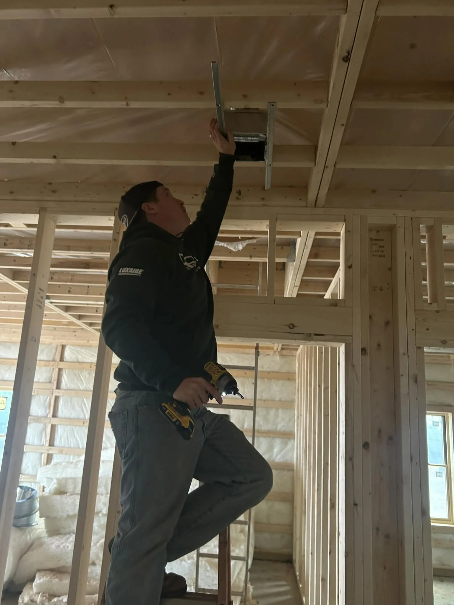 A person working on framing inside a house under construction, using a power drill and reaching up to install or adjust a component on the ceiling.