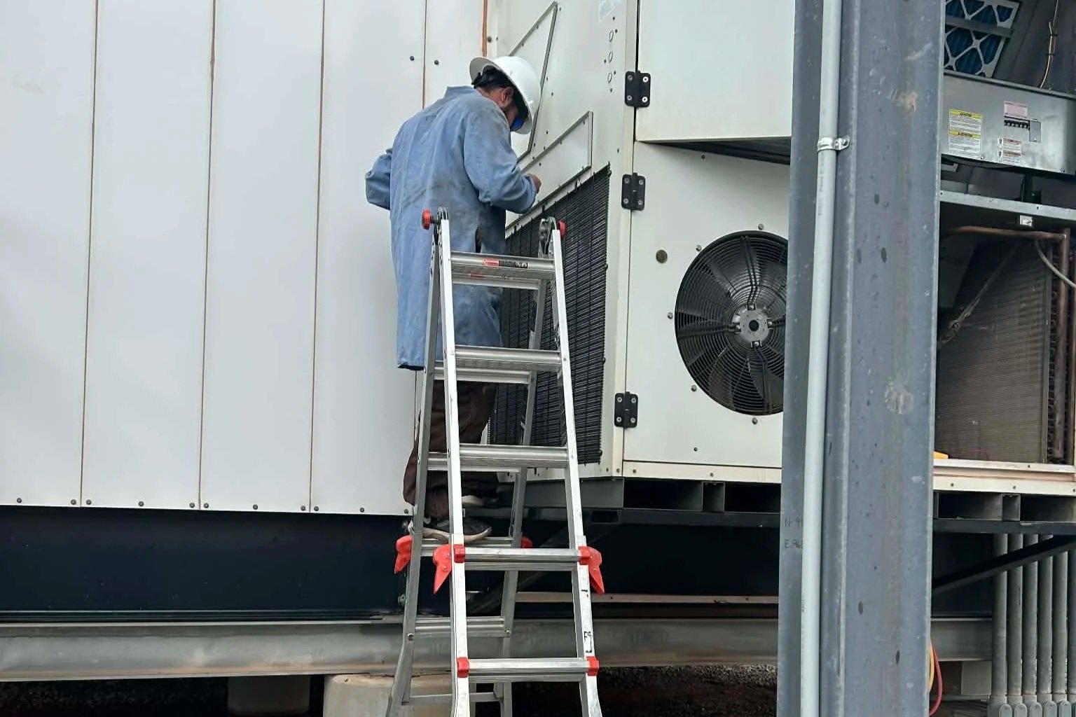 A technician wearing a white protective helmet and blue coveralls working on an industrial HVAC system while standing on a ladder.