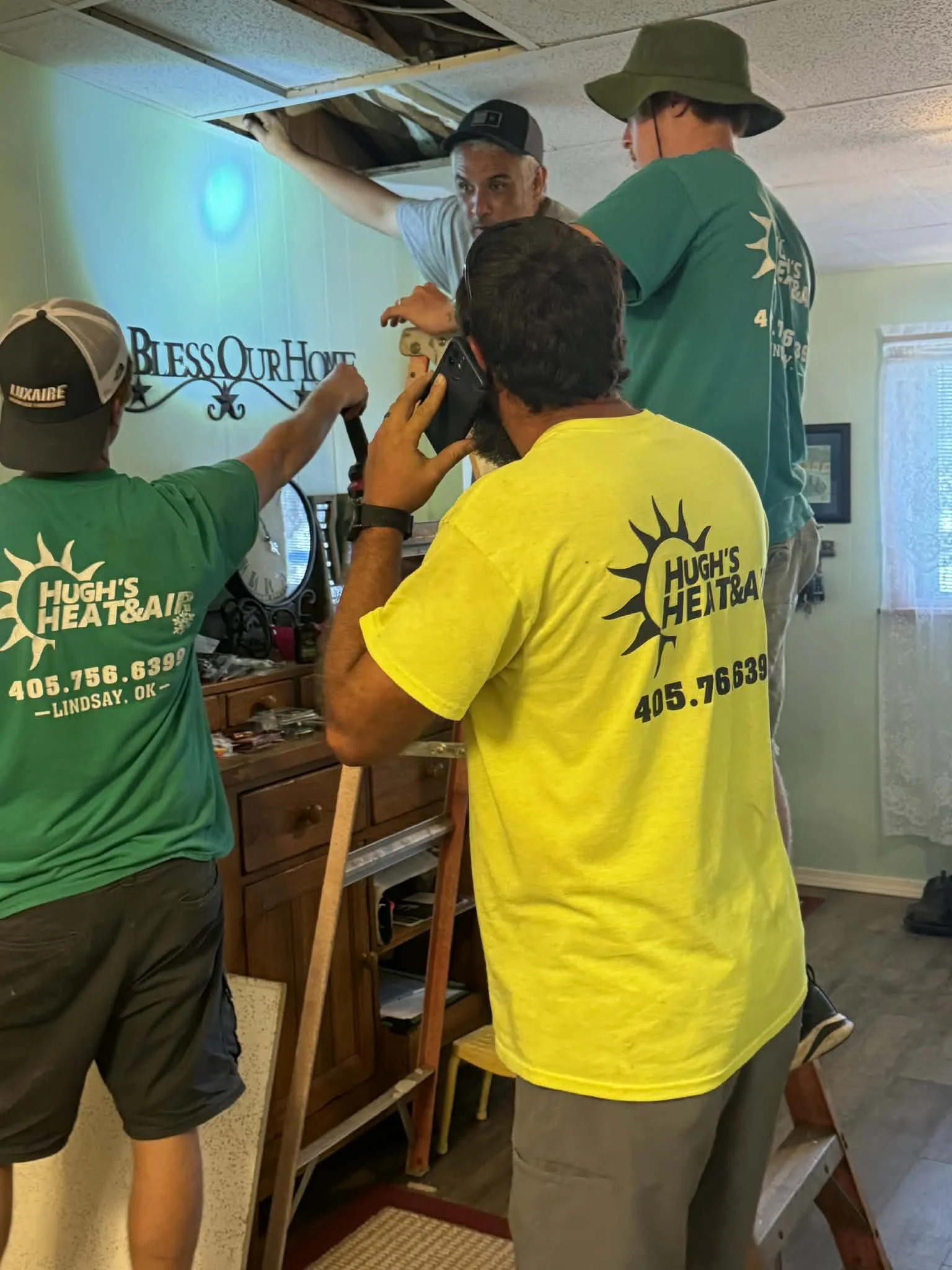 Four men working on ceiling repair in a living room. One man is on a ladder, another is using a hammer, one is on the phone, and the fourth is peering over the others, all engaged in fixing the ceiling.