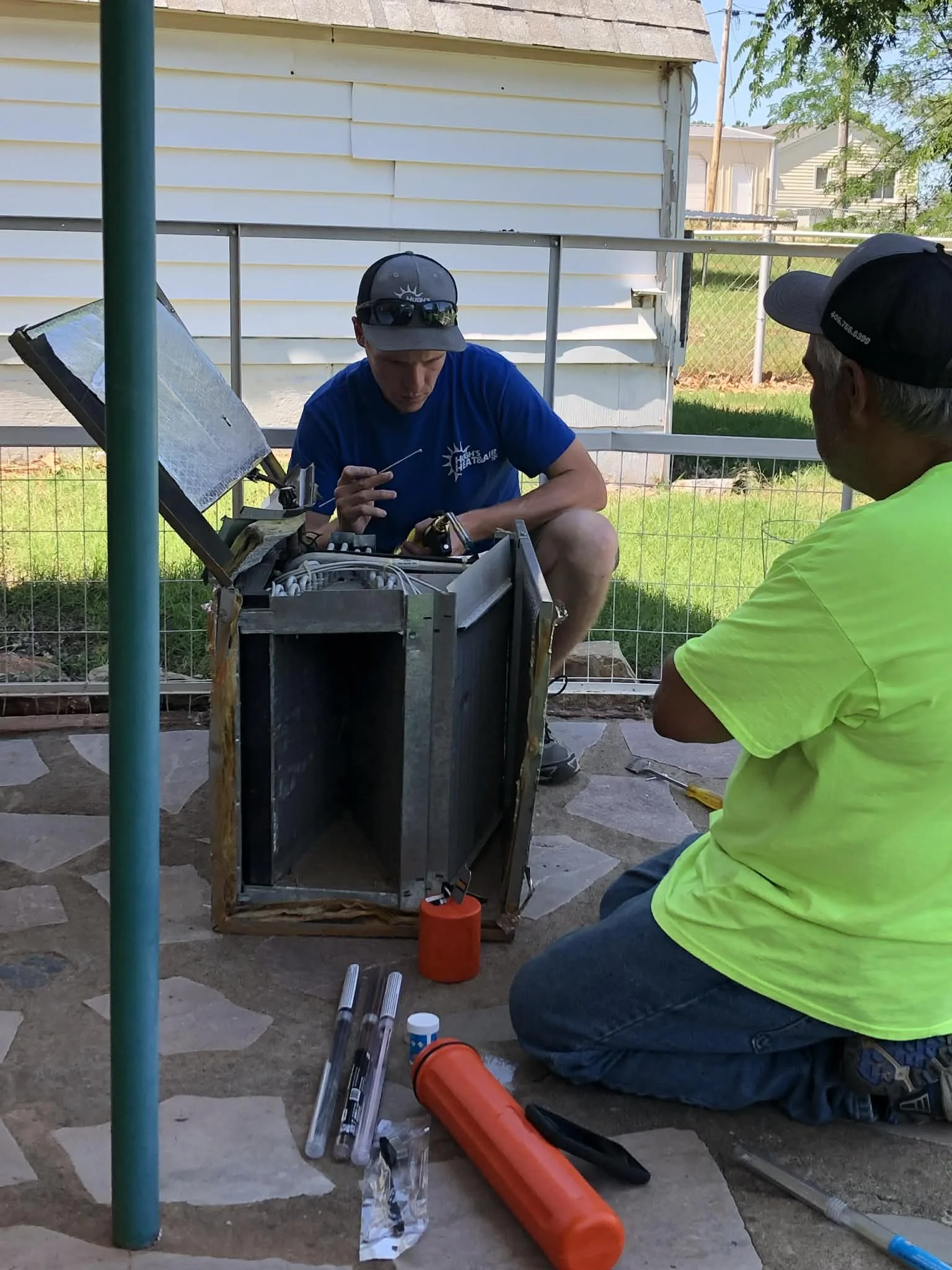 Two men working outdoors on a piece of technical equipment, with tools and parts around them, in a backyard with a white house and green yard.
