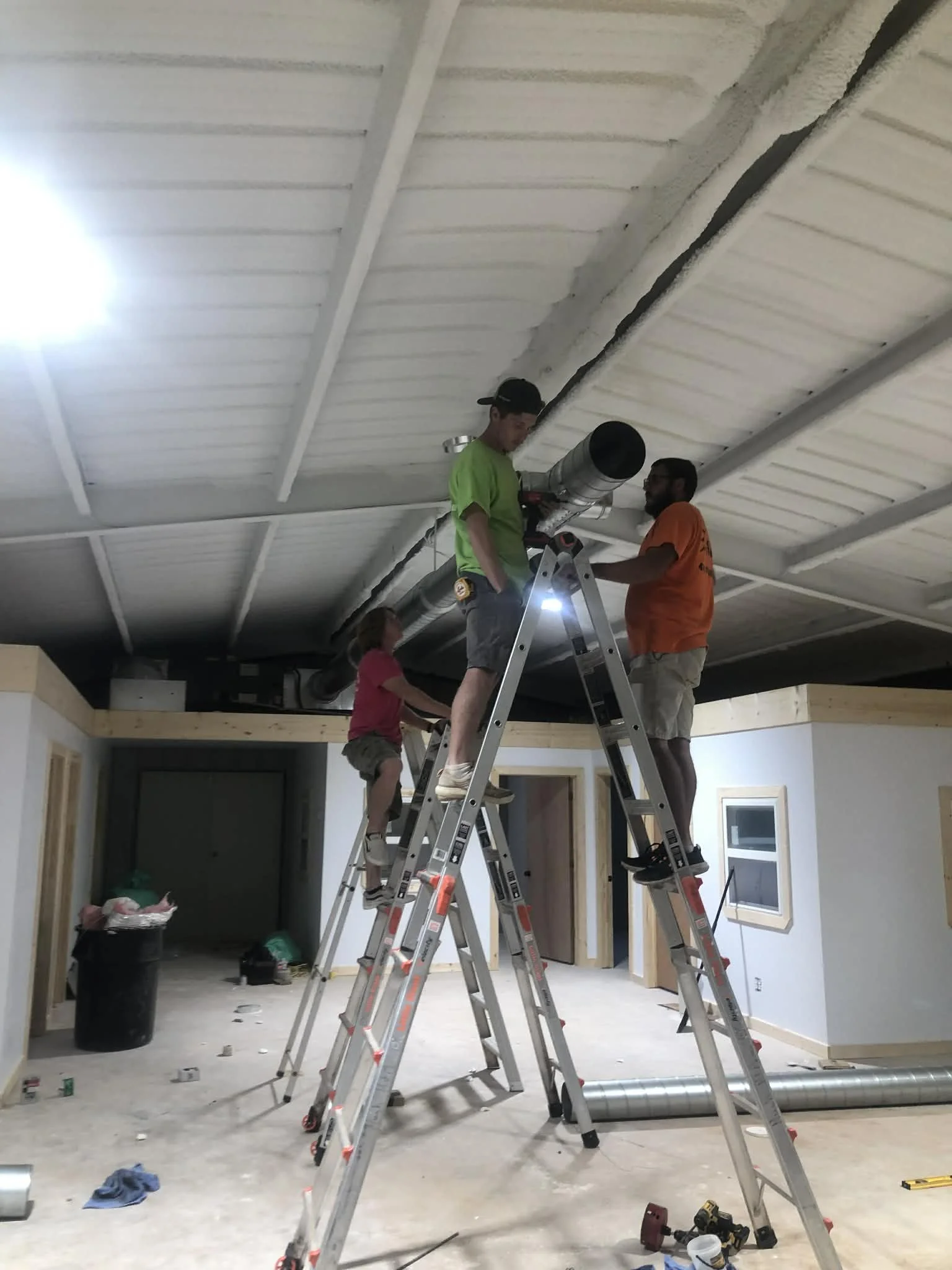 Three people, two adults and a child, installing or repairing ductwork on a ceiling inside a building under construction.
