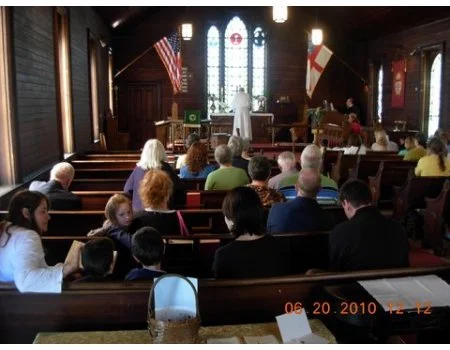 People sitting in pews inside a church with stained glass windows, flags, and an altar at the front.