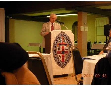 An elderly man in a beige suit speaking at a podium in a conference room. The podium has a seal that reads 'Daughters of North Michigan.' There are tables and chairs, with people attending the event. The timestamp on the photo is 2011, 09:43.