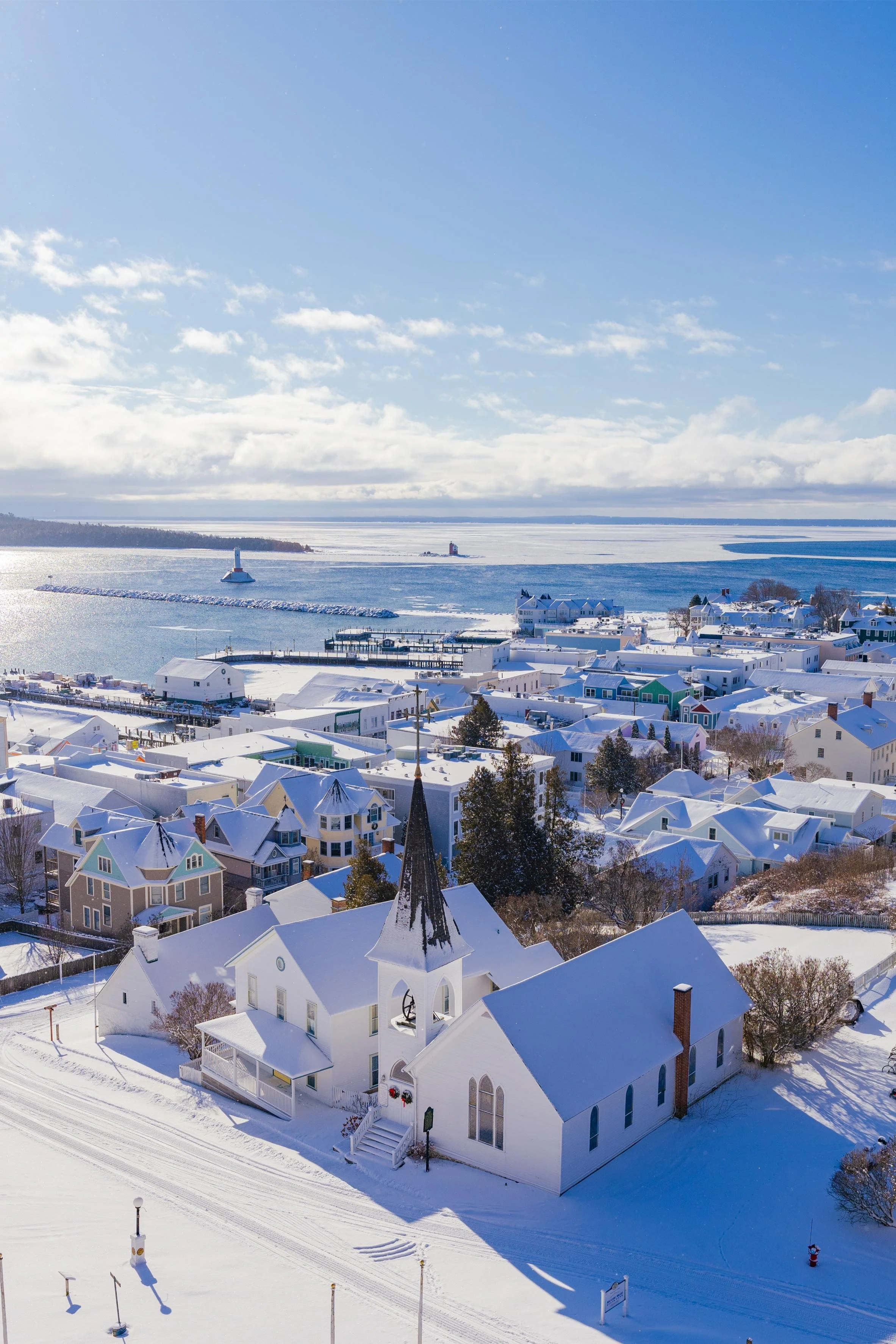 Snow-covered Mackinac Island, Michigan with houses, church, and waterway under blue sky with clouds, winter scene.