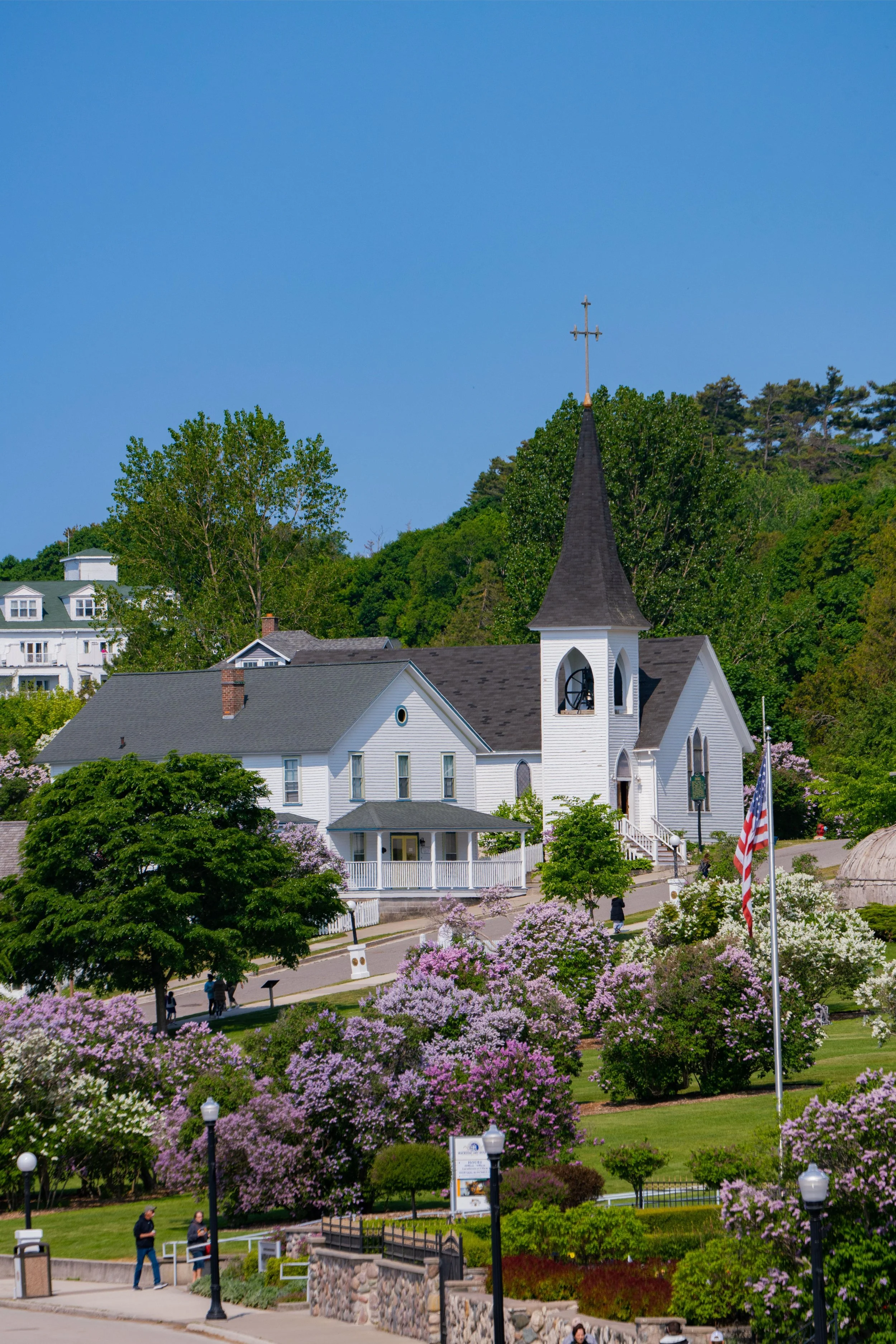 A white church with a tall steeple and cross, surrounded by blooming pink and purple flowers, green trees, and a park with walking paths and people, against a bright blue sky with a hill in the background.
