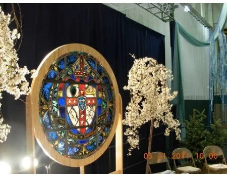 Decorative circular stained glass window with various symbols, surrounded by white flowering trees and green plants, set up indoors.