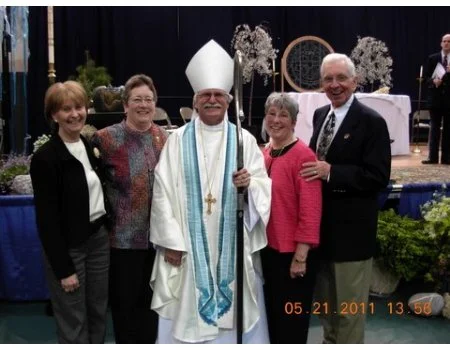 A group of five people, including a man dressed as a bishop or pope, standing together and smiling at a formal event with a decorated stage in the background.