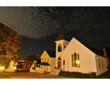 Nighttime view of a white church with a tall steeple illuminated by exterior lights, with trees and other buildings nearby under cloudy sky.
