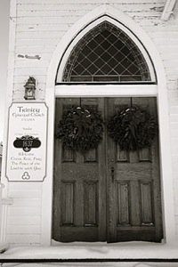 Double wooden door with semi-circular window above, decorated with two hanging wreaths, on a white building with a sign to the left.