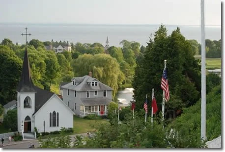 A scenic view of a small town with a white church, a gray house, and several American flags on flagpoles surrounded by green trees and water in the distance.