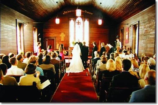 Wedding ceremony taking place inside a wooden church with guests seated facing the altar, where a bride and groom stand before a minister.