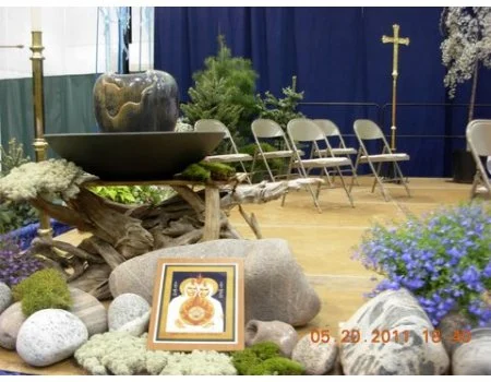 Indoor display with framed religious icon, rocks, flowers, small pine tree, and candles, set up on a wooden stage with blue curtains and chairs in the background.
