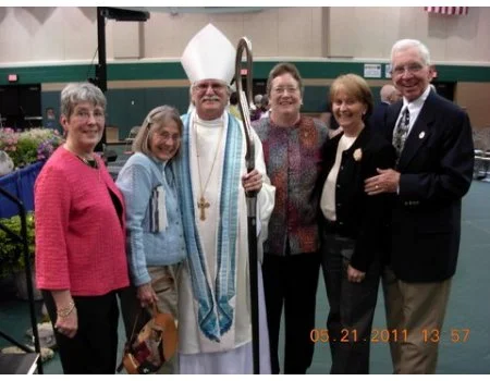 Group of five women and one man, including a bishop in religious robes, standing indoors and smiling at the camera.