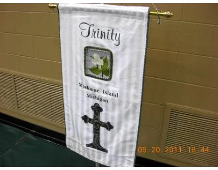 A white church banner with the word 'Trinity' and an image of a poinsettia, located at Maquindna Island, Michigan, hanging on a gold rod against a beige wall.