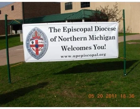 A white banner with the Episcopal Diocese of Northern Michigan logo and text welcoming visitors, set up on a grassy area outside a building.