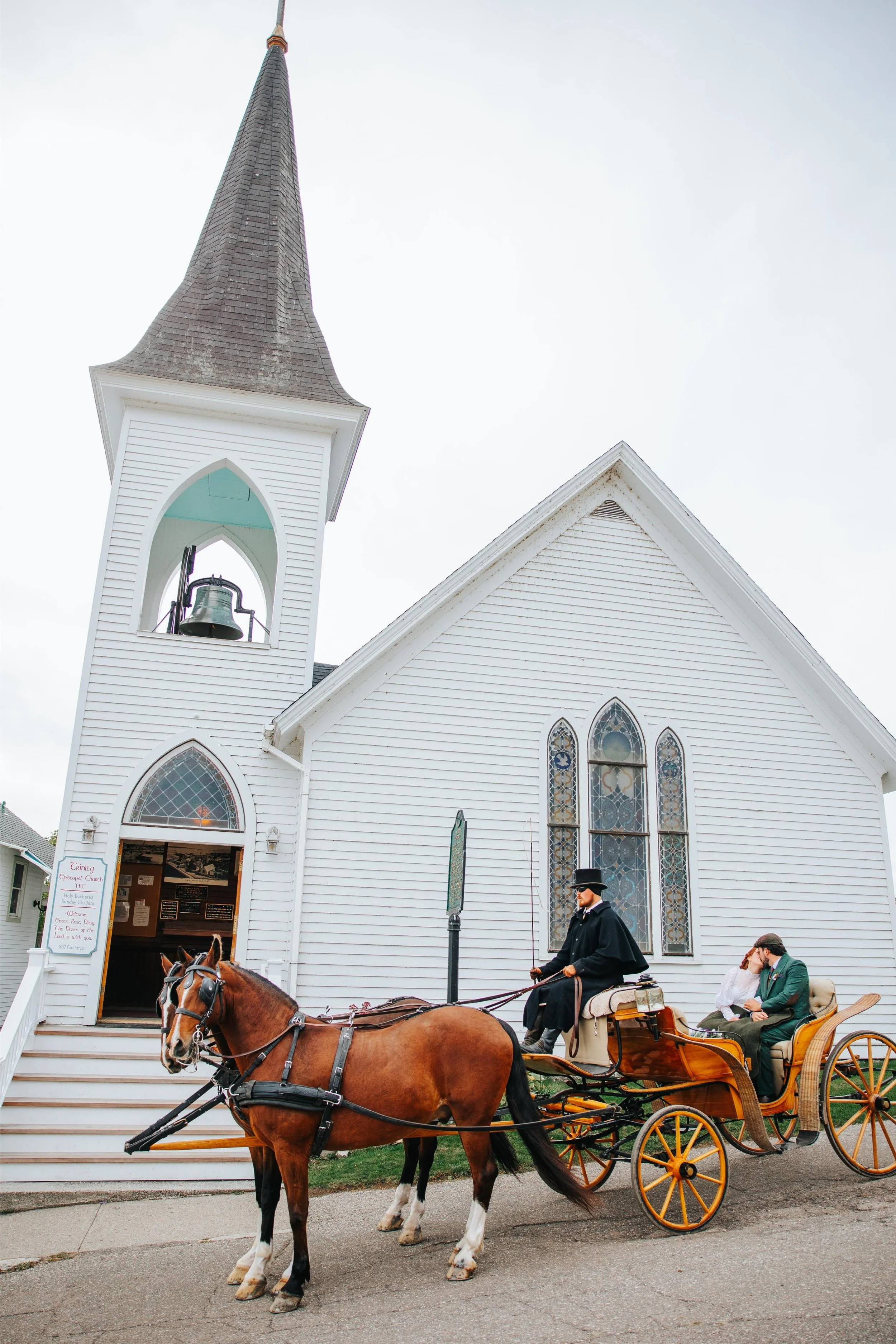 A white church with a tall steeple and stained glass windows. In front, a horse-drawn carriage with two men, one in a uniform and another in casual attire, sitting and talking. The horse is brown with white markings, harnessed to the carriage.