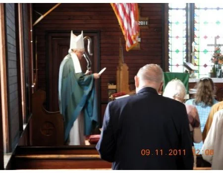 A person dressed as a bishop or priest in a church, reading from a book, with an American flag hanging nearby, and people praying or standing in front of them.