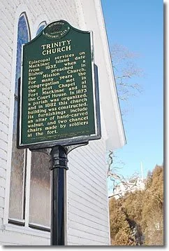 Historical green informational sign for Trinity Church, describing its significance, history, and architecture, mounted on a black pole in front of a building under a clear blue sky.