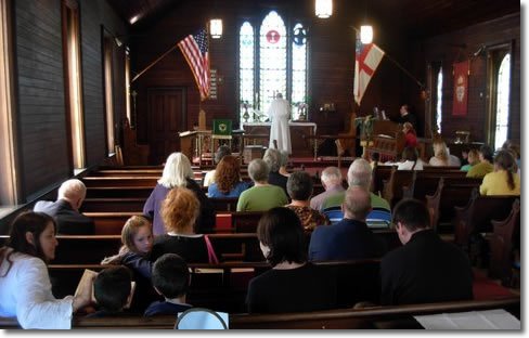 People attending a church service inside a church with wooden walls, stained glass windows, an American flag and a Christian flag, and a cross at the altar.