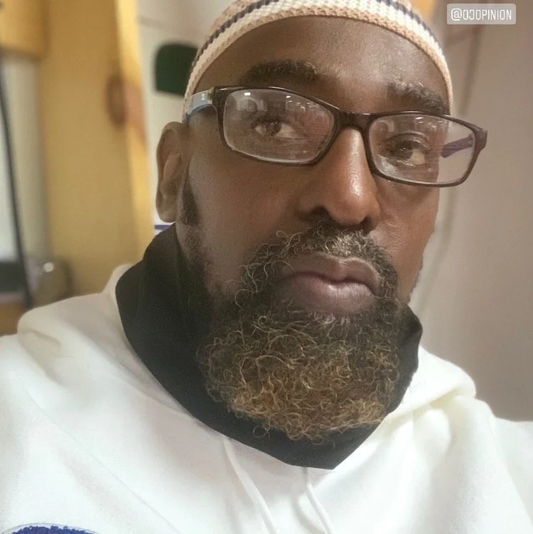 Close-up of a man wearing glasses, a beige cap, a white hoodie, and a black face mask pulled below his chin.