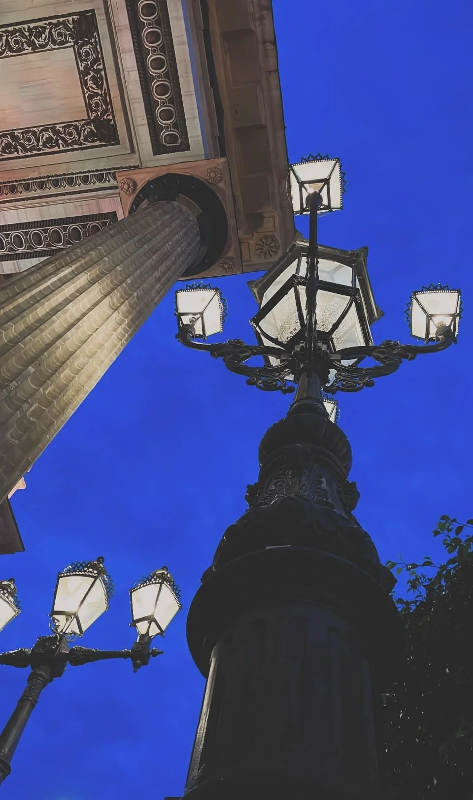 Farola antigua de hierro en una calle, vista desde abajo, contra un cielo azul noche, cerca de un edificio histórico con columnas y detalles decorativos.