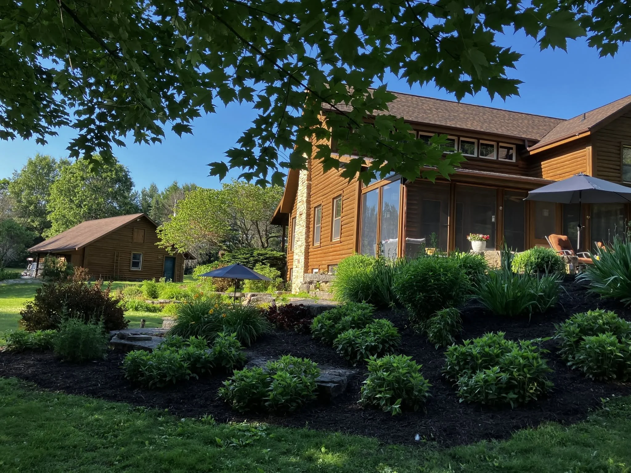 A large wooden house with a patio and umbrellas, surrounded by lush greenery and trees under a clear blue sky.