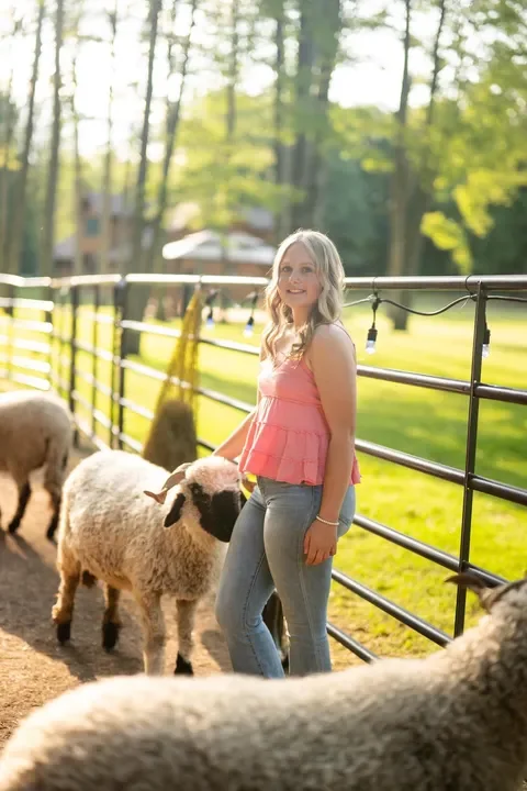 A young woman with long blonde hair in a pink sleeveless top and jeans smiling, standing next to a flock of sheep in an outdoor farm setting, with green trees and a fenced area in the background.