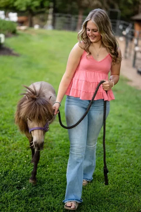 A woman in a pink sleeveless top and jeans walking a small pony on a leash in a grassy outdoor area.