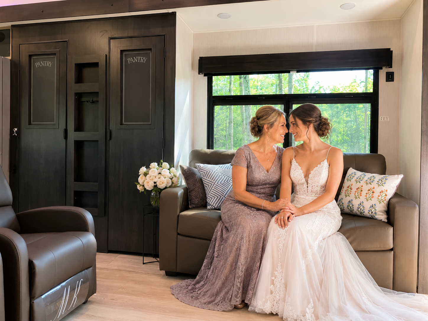 A bride and a woman are sitting on a sofa, smiling at each other, holding hands, with a window showing green trees behind them.