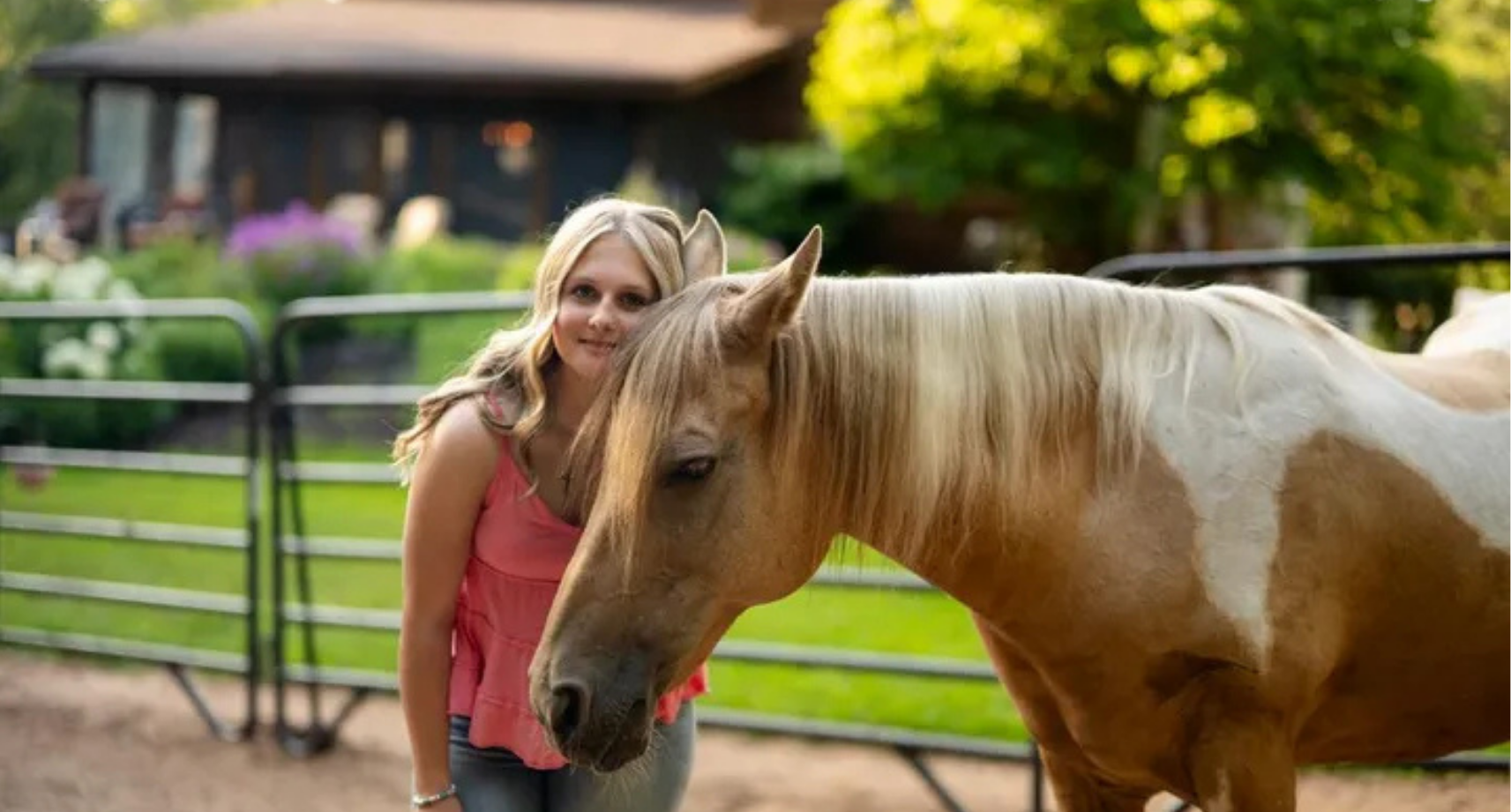 A woman with blonde hair wearing a pink top standing next to a light brown and white horse in a fenced pasture with green trees and a wooden building in the background.
