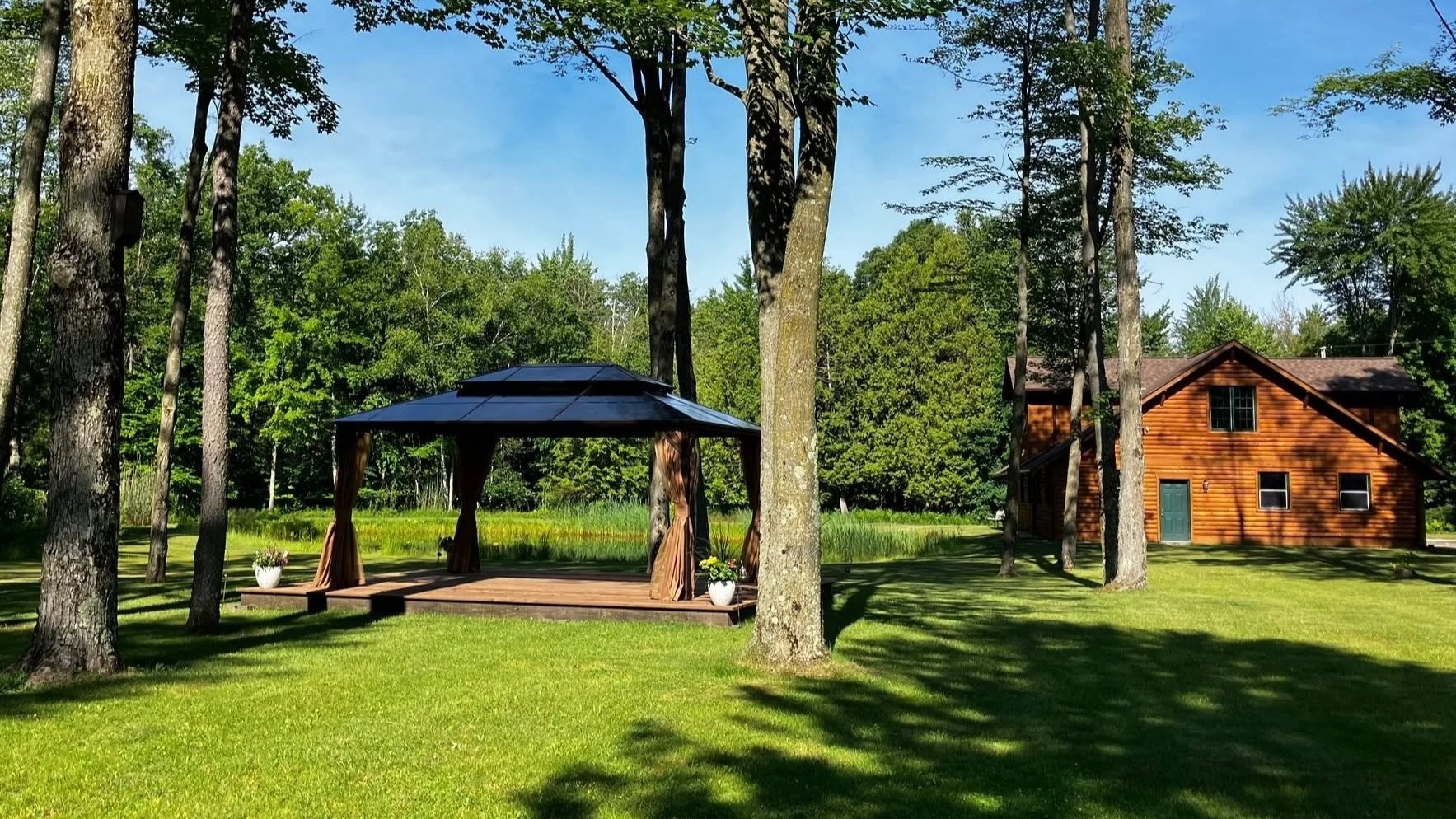 A wooden house with a blue door surrounded by tall trees and a green lawn. There is a small gazebo with curtains and potted plants on a wooden deck.