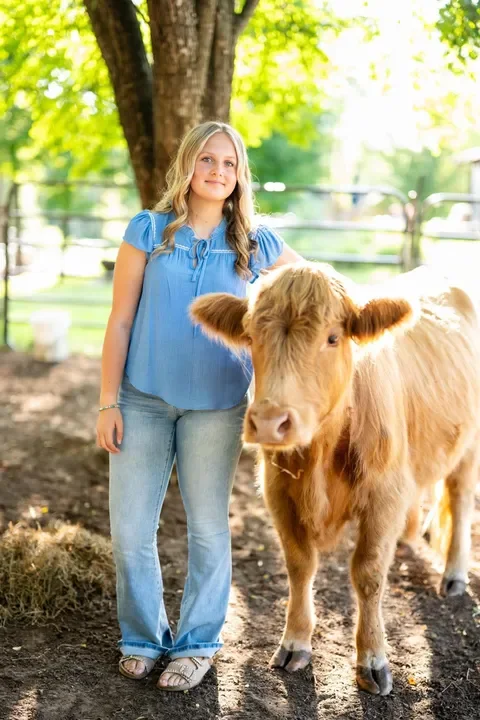 A girl in a blue blouse and jeans standing next to a brown cow in a farm setting with trees and a fence in the background.
