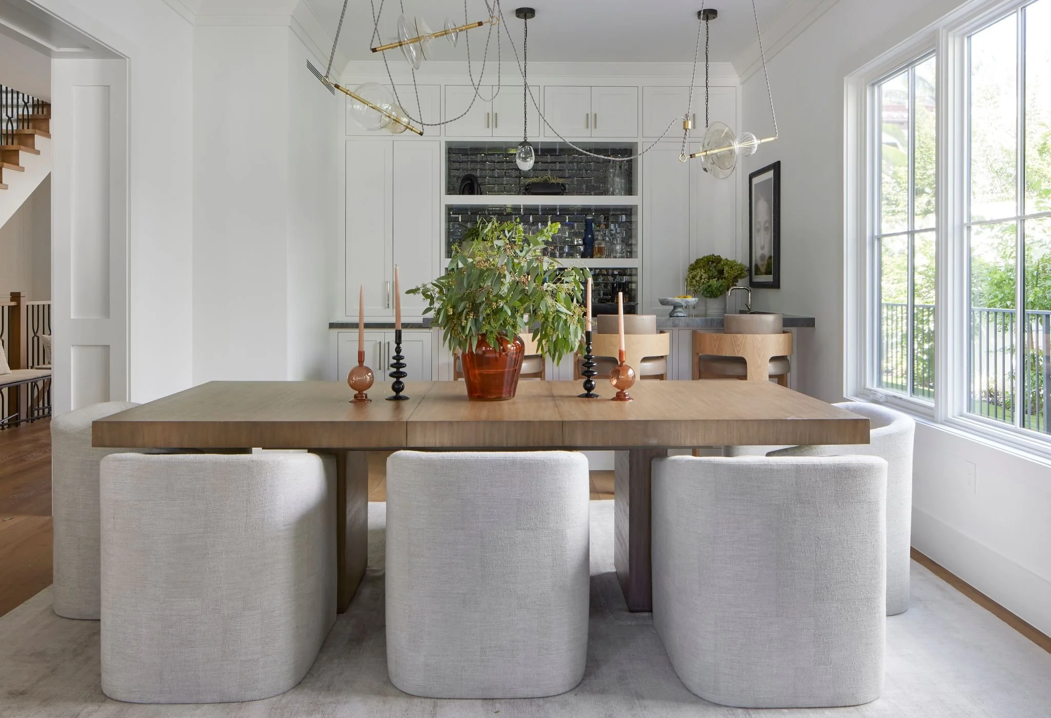 Modern dining room with wooden table, white upholstered chairs, a central vase with green foliage, and candlesticks. Large windows on the right side bring in natural light, and a minimalist chandelier hangs above the table. In the background, there is a small kitchen area with white cabinetry and a window.