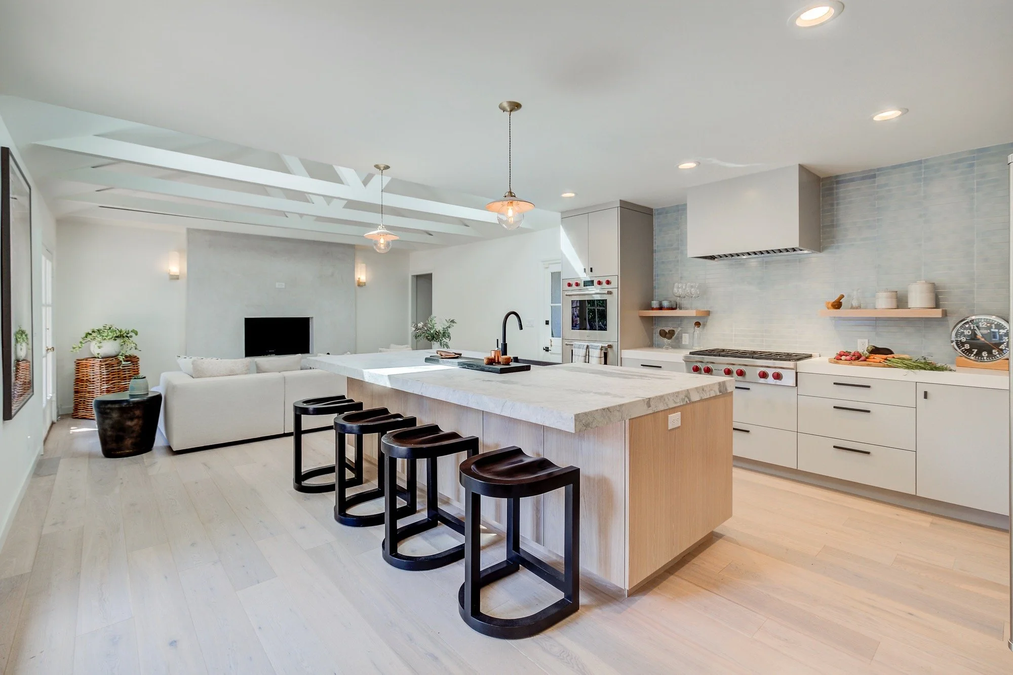 Modern white kitchen with marble island, black bar stools, and open living area with white sofa and TV.
