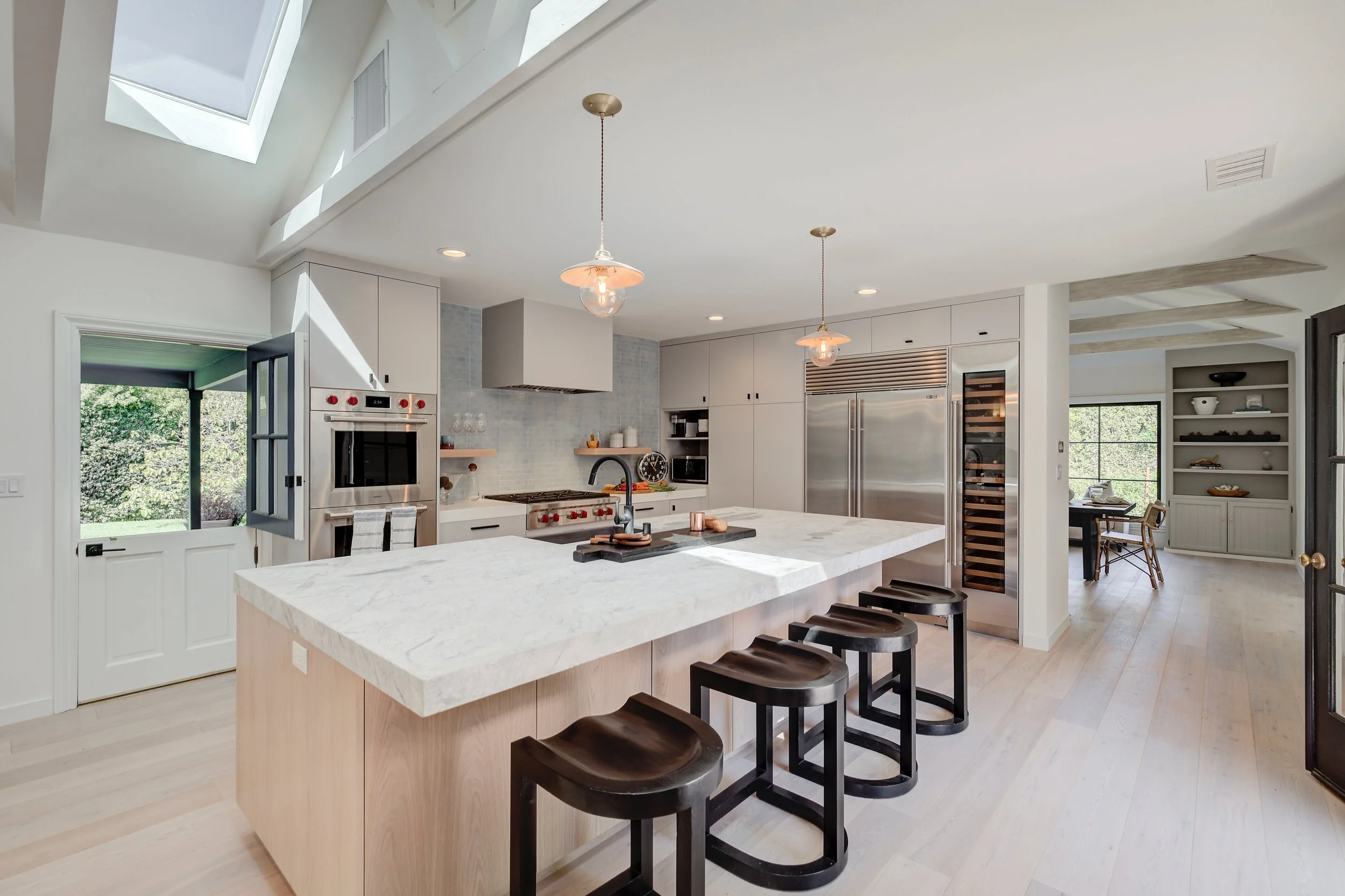 Modern kitchen with white cabinets, marble island, black barstools, stainless steel appliances, and large skylight.